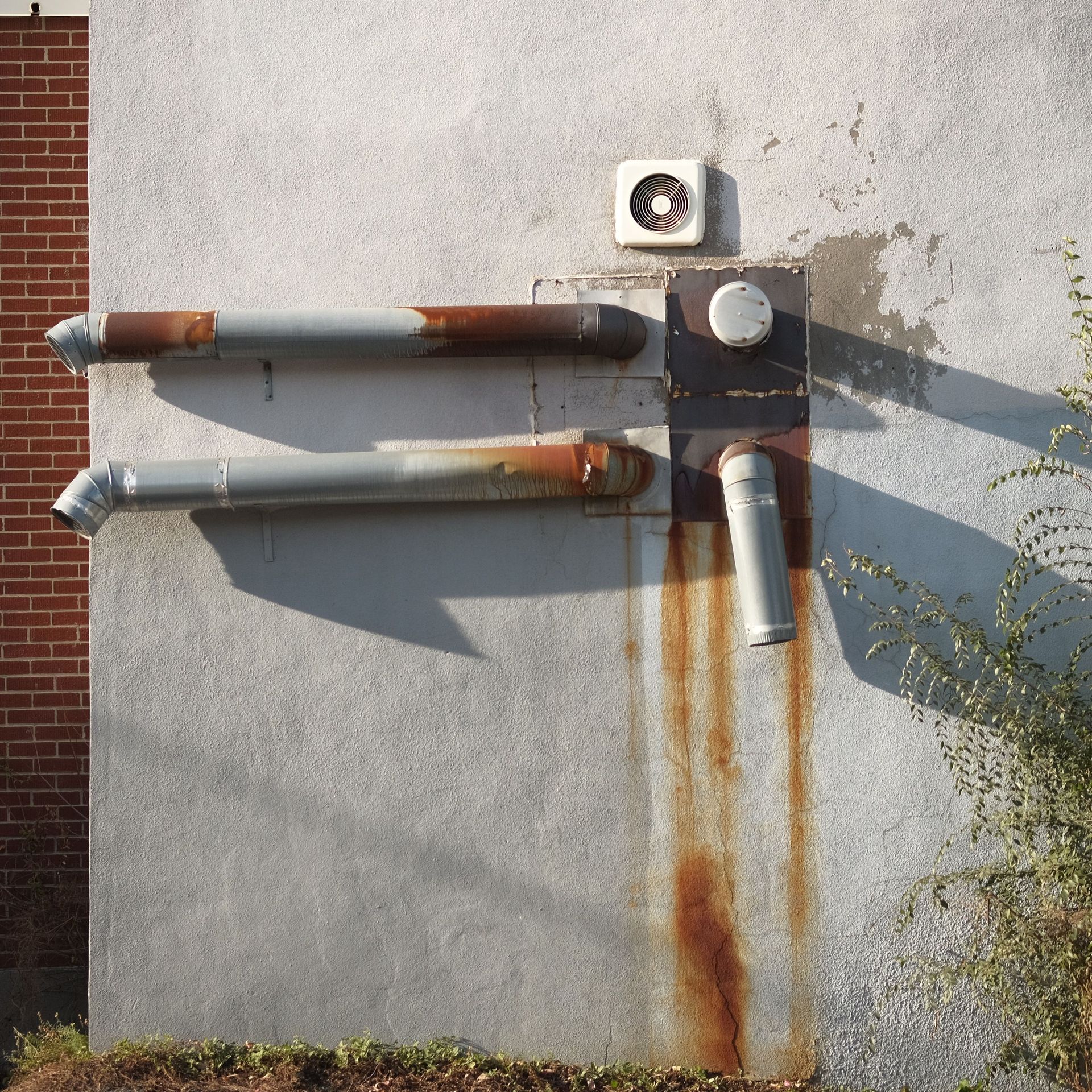 Close-up of rusted pipes and ventilation fan on a concrete wall in sunlight.