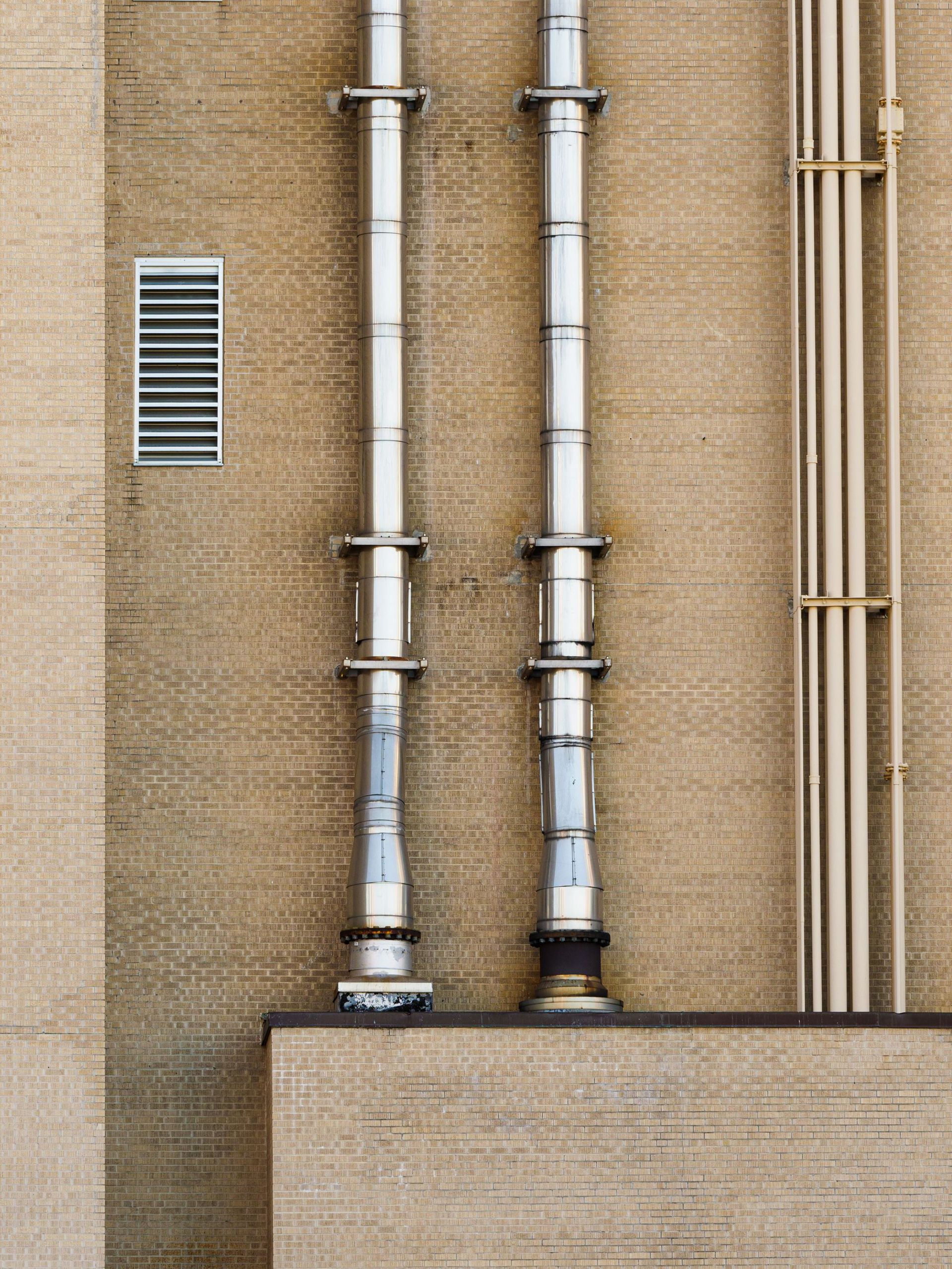 Photo of an industrial building exterior featuring metal pipes and a wall vent.